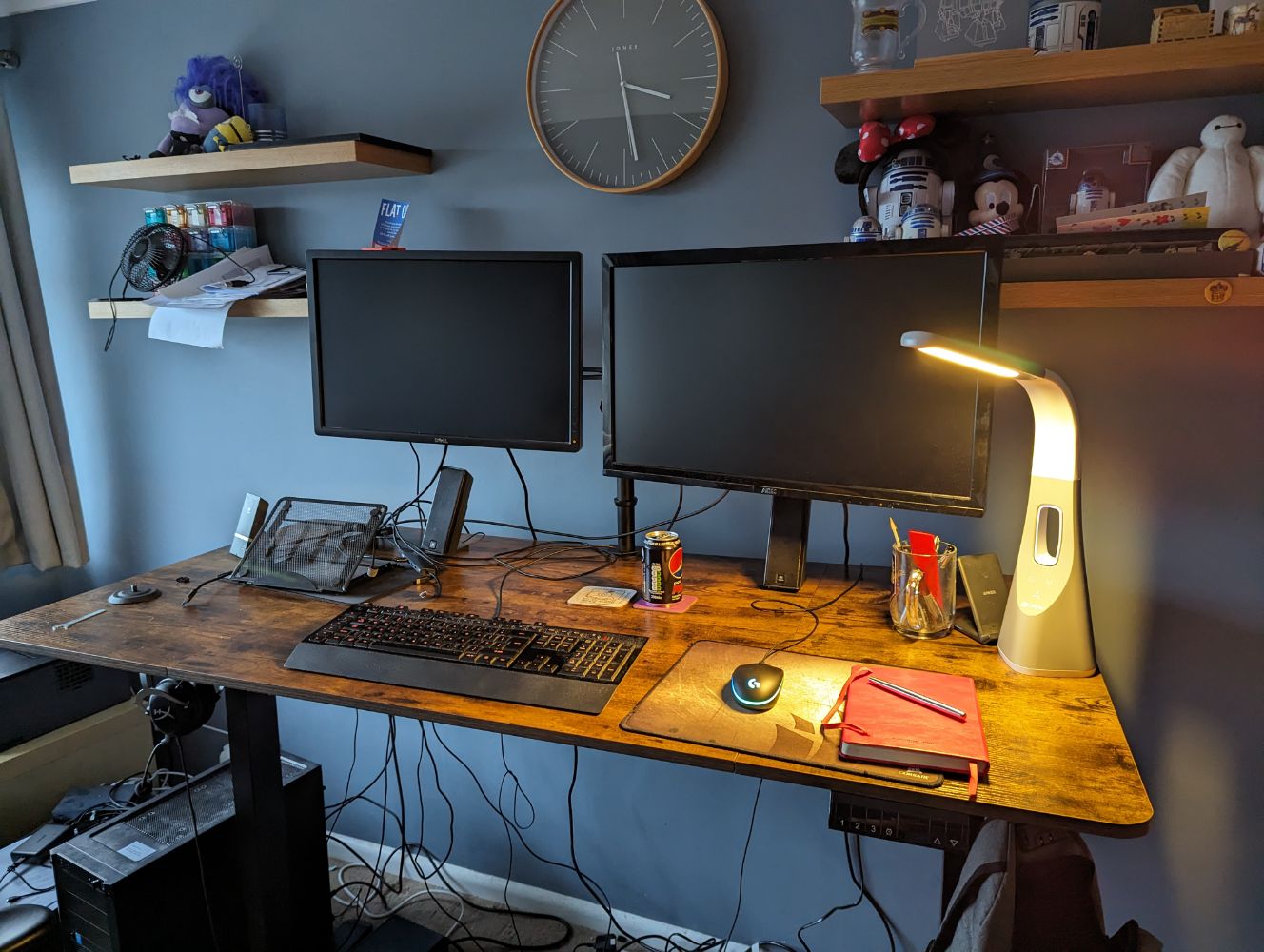 A standing desk with a couple of monitors and a light on it in an office room with blue walls