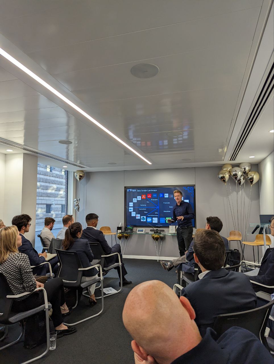 a man stood in front of a presentation being watched by an audience of business people.