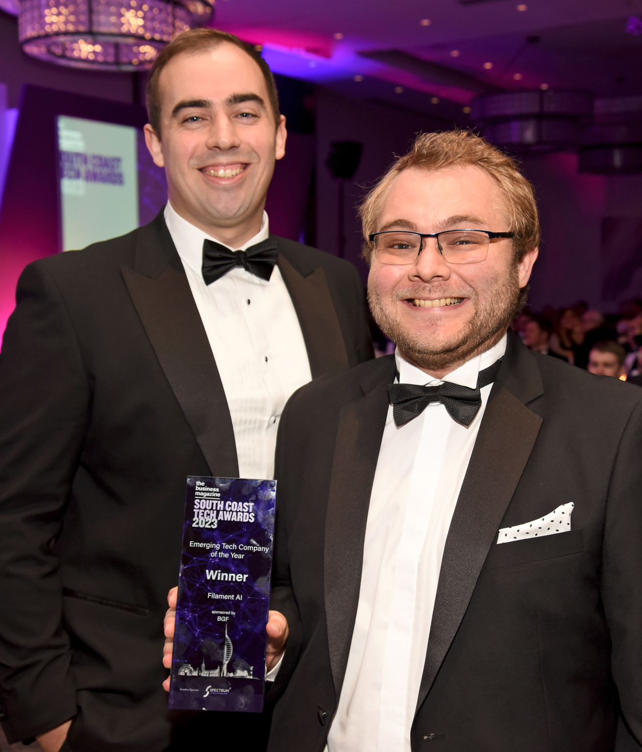 two men wearing tuxedos smiling and holding a glass trophy