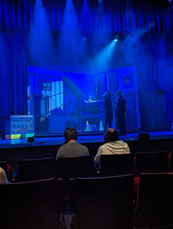 the view of a stage from a theatre auditorium. Two people are stood on the stage under blue lighting with smoke effect