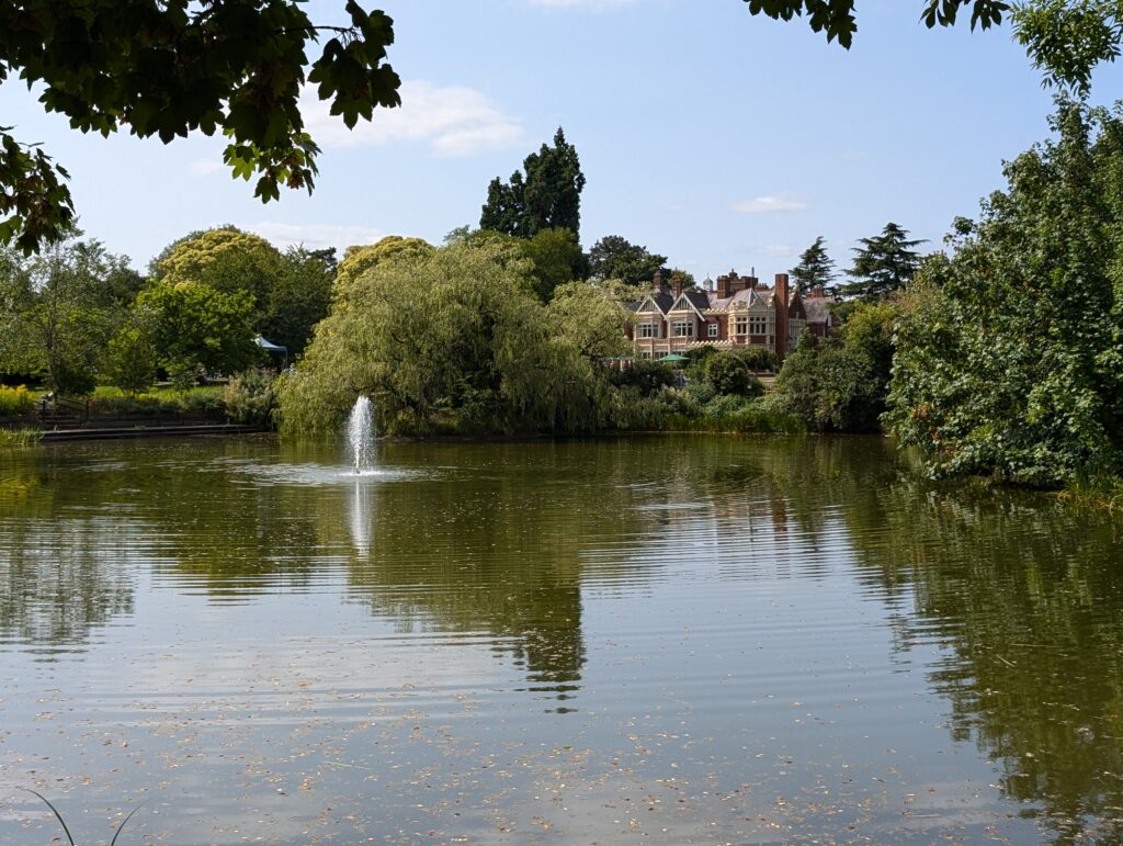 A large house/mansion behind a lake with a fountain at the center of it on a calm, bright and sunny day.