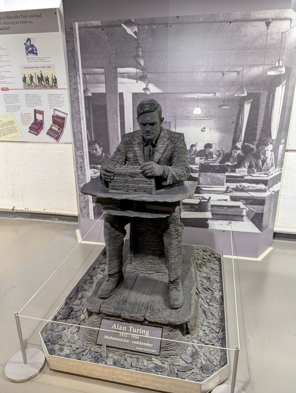 A statue of Alan Turing fashioned from slate set against a black and white photo of an office in Bletchley park