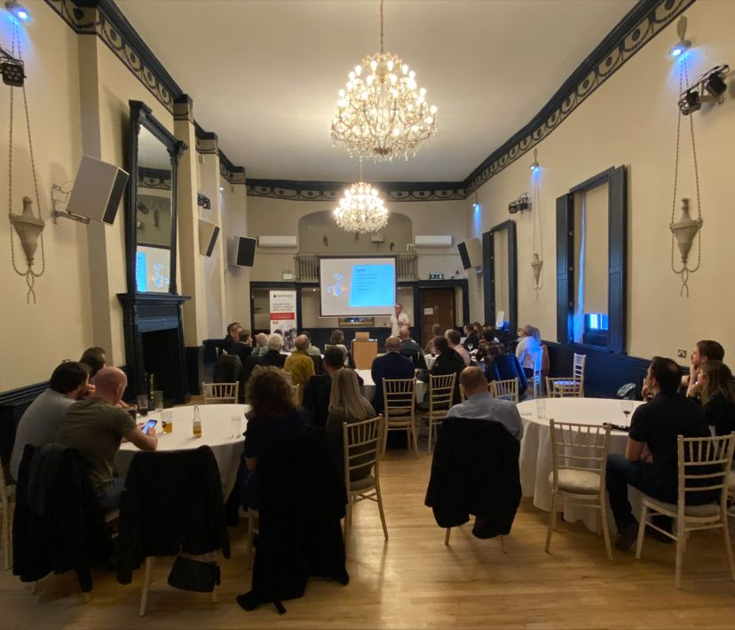 James standing at the front of a function room filled with people siting at round tables.